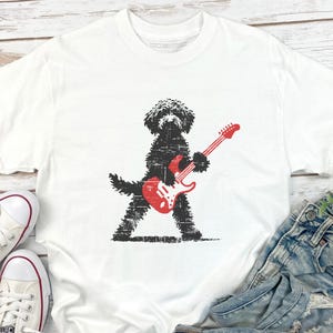 May include: White t-shirt featuring a black dog playing a red electric guitar. The dog is depicted in a distressed, textured style. The guitar has a red body and neck. The shirt is a casual, everyday wear item.
