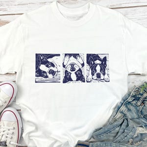 May include: White t-shirt featuring a navy blue graphic of three Boston Terriers in different poses. The dogs are in a rectangular design. The shirt is paired with blue jeans and white sneakers.