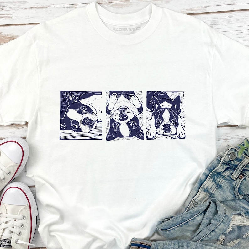 May include: White t-shirt featuring a navy blue graphic of three Boston Terriers in different poses. The dogs are in a rectangular design. The shirt is paired with blue jeans and white sneakers.