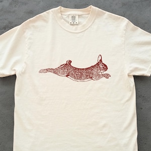 May include: Off-white t-shirt featuring a detailed illustration of a running rabbit in a reddish-brown hue. The design is centered on the shirt, showcasing the animal's form. The shirt has short sleeves and a crew neck.