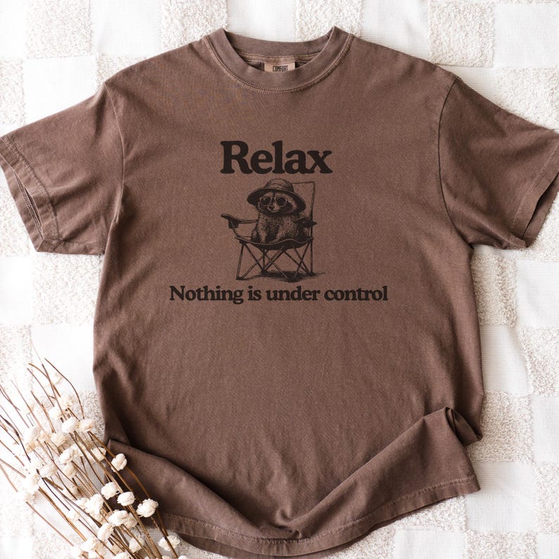 May include: Brown t-shirt with the word "Relax" above an illustration of an animal wearing a hat and sunglasses, sitting in a folding chair. The text "Nothing is under control" is below the illustration.
