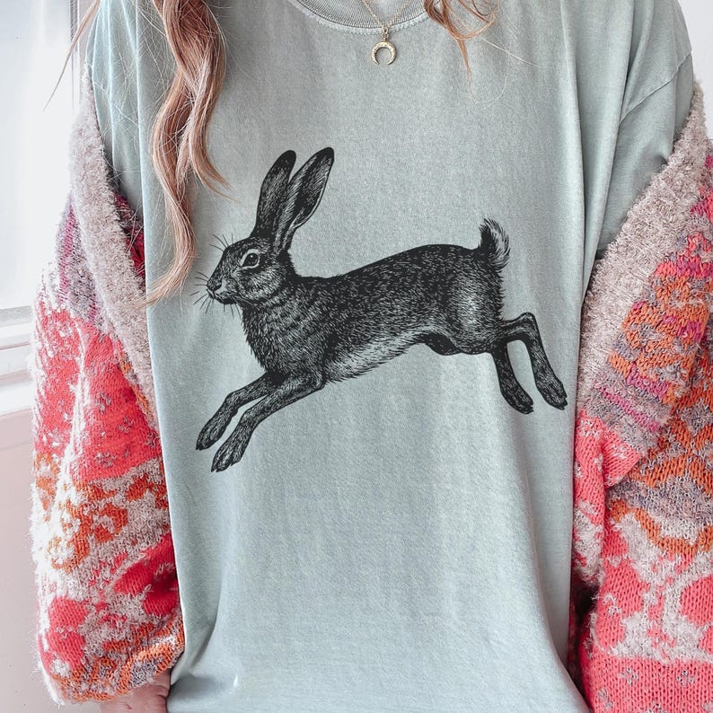May include: A light blue t-shirt featuring a detailed black and white illustration of a running hare. The graphic is centered on the shirt. The person is wearing a pink and orange patterned cardigan.