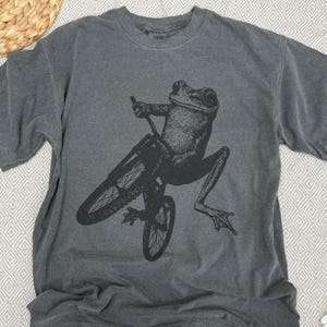 Pu&ograve; includere: T-shirt grigio scuro con una grafica nera di una rana in bicicletta. La rana &egrave; a mezz'aria, con le gambe distese. La bicicletta &egrave; nera, con raggi e manubrio dettagliati. La maglietta ha un girocollo e maniche corte.