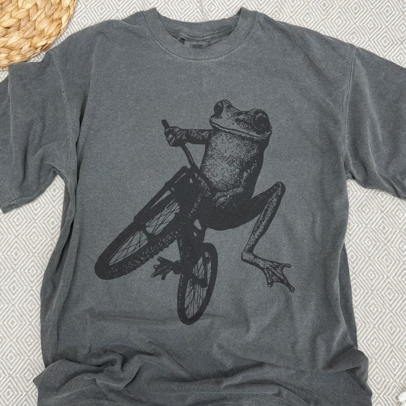 Pu&ograve; includere: T-shirt grigio scuro con una grafica nera di una rana in bicicletta. La rana &egrave; a mezz'aria, con le gambe distese. La bicicletta &egrave; nera, con raggi e manubrio dettagliati. La maglietta ha un girocollo e maniche corte.