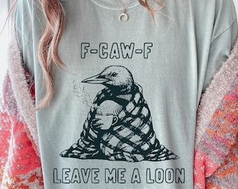 May include: A light blue t-shirt with a graphic of a loon wrapped in a blanket, holding a coffee cup. The text "F-CAW-F" is above the graphic, and "LEAVE ME A LOON" is below.