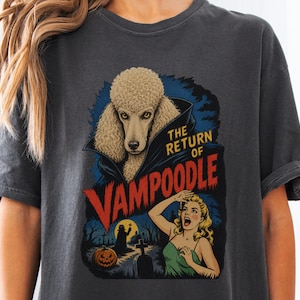 May include: A charcoal grey t-shirt featuring a graphic print of a poodle with a vampire cape, titled THE RETURN OF VAMPOODLE in red and yellow text. The illustration includes a frightened person, a pumpkin, and a graveyard scene at night.
