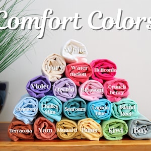 Pu&ograve; includere: Un'esposizione di magliette arrotolate in vari colori, tra cui bianco, viola e blu laguna. Il testo "Comfort Colors" &egrave; visualizzato in alto, con nomi di colori come "Orchid" e "Kiwi" che etichettano ogni maglietta.