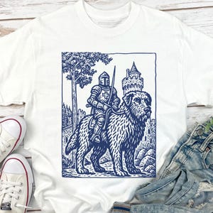 May include: White t-shirt featuring a blue illustration of a knight riding a large dog, with a castle and tree in the background. The graphic has a vintage, woodcut style. The shirt is paired with denim jeans and white sneakers.