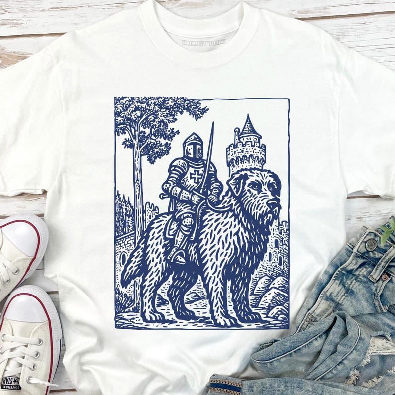 May include: White t-shirt featuring a blue illustration of a knight riding a large dog, with a castle and tree in the background. The graphic has a vintage, woodcut style. The shirt is paired with denim jeans and white sneakers.