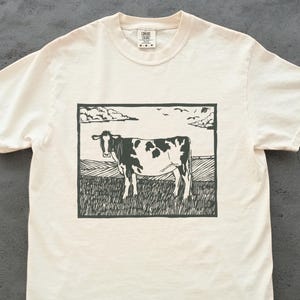 May include: Cream-colored t-shirt featuring a black and white illustration of a cow standing in a field. The cow is the focal point, set against a backdrop of a rural landscape with a sky and clouds. The shirt has a crew neck and short sleeves.