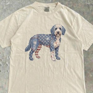 May include: A cream-colored t-shirt featuring a graphic of a dog with a patriotic design. The dog's fur is patterned with blue denim, white stars, and red and white stripes, evoking an American flag theme.