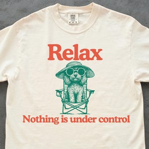 May include: Cream-colored t-shirt with the word "Relax" in orange above a green illustration of a dog wearing sunglasses and a hat, seated in a beach chair. The text "Nothing is under control" is below the dog.