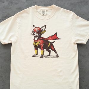 以下が含まれることがあります： クリーム色のTシャツ。スーパーヒーローのチワワのイラストが描かれています。犬は赤いと黄色のスーパーヒーローのコスチュームを着ており、マントと翼のアクセントが付いています。グラフィックはシャツの中央に配置されています。