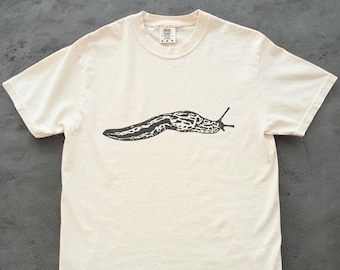 May include: Cream-colored t-shirt featuring a black graphic of a slug. The slug illustration is detailed with a patterned body and antennae. The shirt has short sleeves and a crew neck, suitable for casual wear.