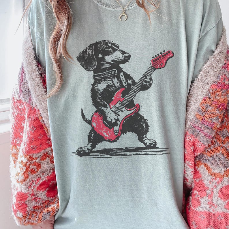 K&ouml;nnte beinhalten: Hellblaues T-Shirt mit einer schwarz-roten Grafik eines Dackels, der eine rote E-Gitarre spielt. Der Hund tr&auml;gt ein Halsband. Das Shirt ist ein l&auml;ssiger Stil.
