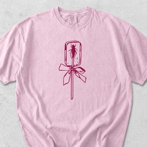 May include: A light pink t-shirt featuring a maroon graphic of a lollipop with an insect inside. The lollipop has a bow and stick. The shirt has a crew neck and short sleeves.