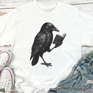 May include: White t-shirt featuring a black illustration of a raven reading a book. The raven is detailed with textured feathers and is holding a small, open book. The shirt is made of a soft material and has a classic crew neck.