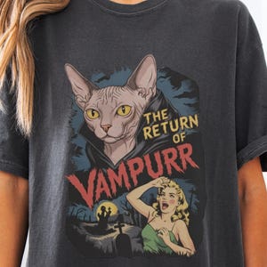 May include: Dark gray t-shirt featuring a graphic of a Sphynx cat in a vampire cape with the text "THE RETURN OF VAMPURR". The design includes a screaming woman and a graveyard scene, creating a vintage horror movie aesthetic.