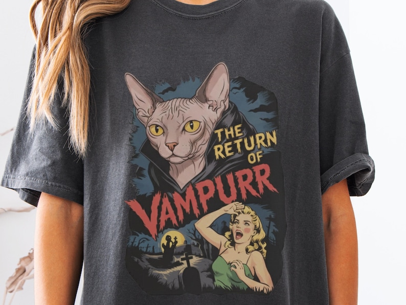 Peut inclure: T-shirt gris fonc&eacute; avec un motif d'un chat Sphynx en cape de vampire et le texte "THE RETURN OF VAMPURR". Le design comprend une femme hurlante et une sc&egrave;ne de cimeti&egrave;re, &eacute;voquant un style de film d'horreur vintage.