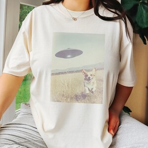May include: A white t-shirt with a graphic print of a corgi running towards a flying saucer. The graphic is in a vintage style.