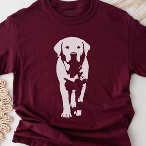 May include: Burgundy t-shirt featuring a white graphic of a Labrador Retriever dog. The dog is depicted in a simple, minimalist style, walking forward. The shirt has a crew neck and short sleeves.