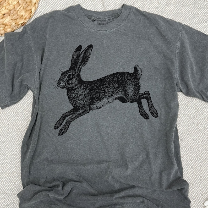 May include: Gray t-shirt featuring a black illustration of a leaping hare. The graphic is detailed, showing the hare's fur and posture. The shirt has a classic crew neck and short sleeves, suitable for casual wear. The fabric appears soft and pre-washed.