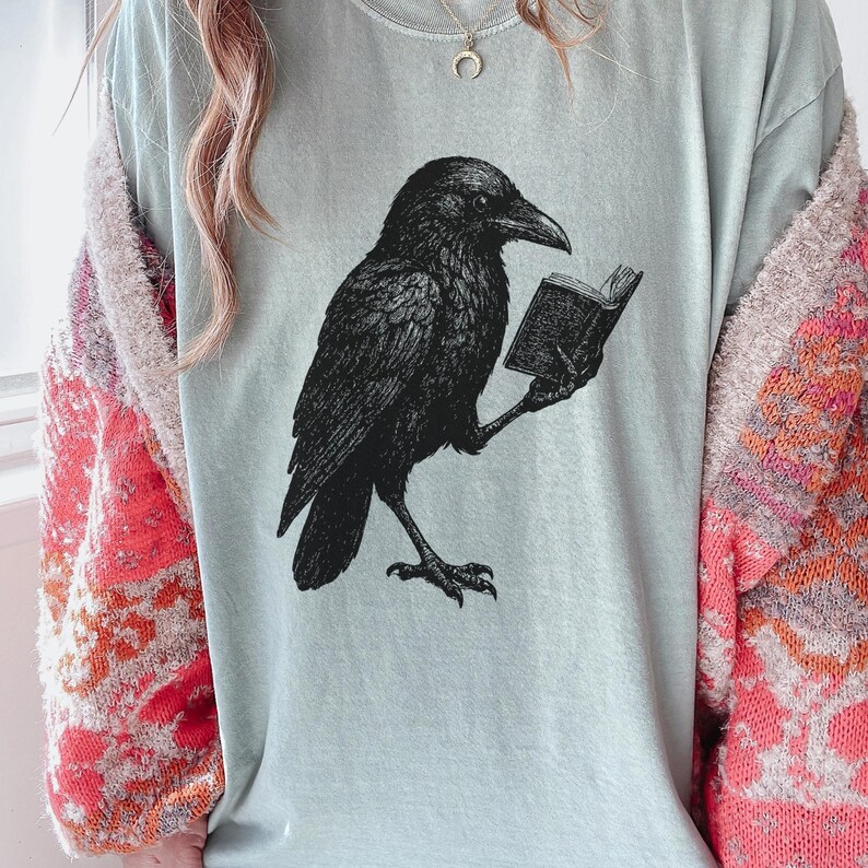 May include: A light blue t-shirt featuring a black illustration of a raven reading a book. The raven is detailed with textured feathers. The shirt is a comfortable fit, suitable for casual wear.