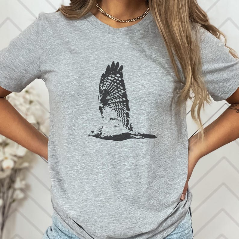 May include: A heather gray t-shirt featuring a detailed black and white illustration of a bird in flight. The bird's wings are spread, showcasing intricate feather patterns. The shirt has a classic crew neck and short sleeves.