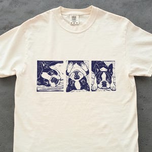 May include: Cream-colored t-shirt featuring a navy blue linocut print of three Boston Terriers in different poses. The dogs are depicted in rectangular frames. The shirt has a crew neck and short sleeves.