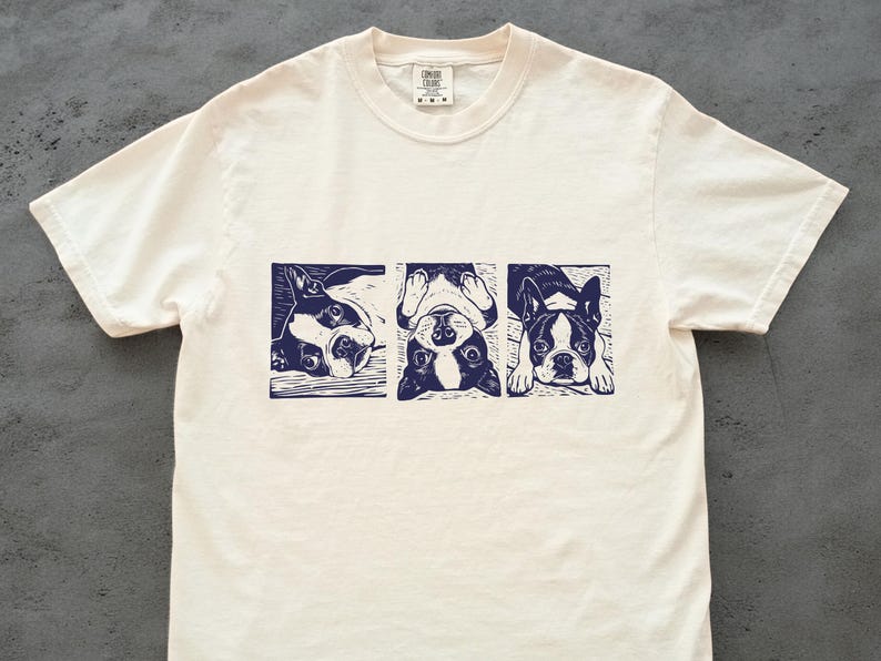 May include: Cream-colored t-shirt featuring a navy blue linocut print of three Boston Terriers in different poses. The dogs are depicted in rectangular frames. The shirt has a crew neck and short sleeves.