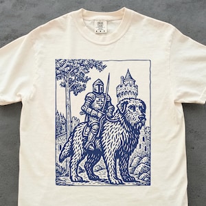 May include: Off-white t-shirt featuring a navy blue illustration of a knight in armor riding a large dog, with a castle and tree in the background. The design is a detailed, vintage-style print.