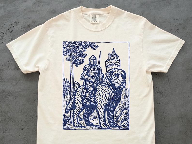 May include: Off-white t-shirt featuring a navy blue illustration of a knight in armor riding a large dog, with a castle and tree in the background. The design is a detailed, vintage-style print.