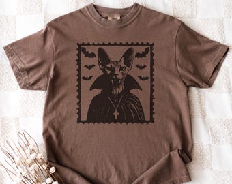 Puede incluir: Camiseta marrón con un gráfico negro de un gato vampiro en un diseño de sello postal. El gato lleva una capa y un collar con una cruz, con murciélagos en el fondo. La camiseta es de material suave.