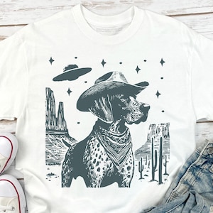 Puede incluir: Camiseta blanca con un perro con sombrero de vaquero y pañuelo, sobre un paisaje desértico con un OVNI. El diseño es en tonos grises y negros, con estrellas y cactus. Se ven zapatillas blancas y vaqueros.