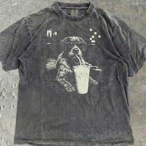 May include: A charcoal gray t-shirt featuring a graphic of a dog wearing sunglasses and drinking from a cup with a straw. The dog is sitting in a chair. The shirt has a vintage, washed appearance.