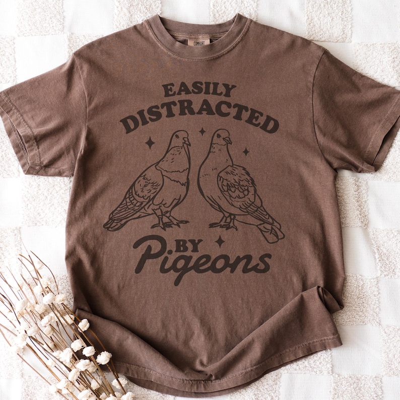 May include: A brown t-shirt featuring the text "EASILY DISTRACTED BY PIGEONS" and a graphic of two pigeons. The shirt is a solid brown colour and has short sleeves.