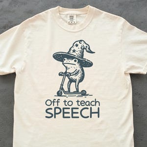 Op de afbeelding: Crèmekleurig T-shirt met een donkerblauwe illustratie van een kikker met een tovenaars hoed op een scooter. De tekst "Off to teach SPEECH" staat eronder. Een casual kledingstuk.