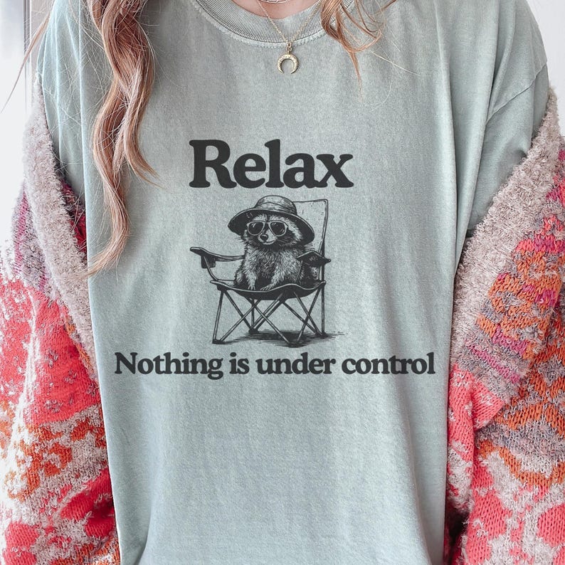 May include: A light blue t-shirt with the word "Relax" above a graphic of a raccoon wearing sunglasses and a hat, seated in a folding chair. The text "Nothing is under control" is below the graphic.
