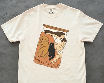 Könnte beinhalten: Cremefarbenes T-Shirt mit einer Grafik eines Bettes mit einer Person, einem Hund und einer Katze. Das Wort "Petriarchy" ist in brauner Schrift auf der Bettdecke aufgedruckt. Das Shirt hat kurze Ärmel und einen Rundhalsausschnitt.