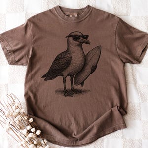K&ouml;nnte beinhalten: Ein braunes T-Shirt mit kurzen &Auml;rmeln und einer schwarzen Grafik im Tintenstil, die eine M&ouml;we mit Sonnenbrille zeigt, die ein Surfbrett unter dem Fl&uuml;gel h&auml;lt. Das Motiv ist mittig auf der Brust des Kleidungsst&uuml;cks mit Rundhalsausschnitt platziert.