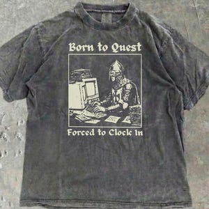 Puede incluir: Camiseta gris oscuro con un gráfico de un caballero en un ordenador, con el texto "Born to Quest" y "Forced to Clock In". La camiseta tiene un aspecto vintage lavado.