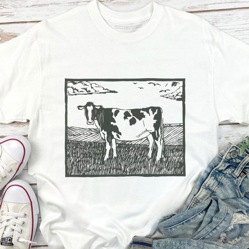 May include: White t-shirt featuring a black and white illustration of a cow standing in a field. The cow has black spots and is set against a background of a field, sky, and clouds. The shirt is paired with denim jeans and white sneakers.
