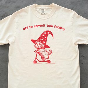 Op de afbeelding: Crèmekleurig T-shirt met een rode afbeelding van een hond met een tovenaars hoed op een scooter. De tekst "off to commit tom foolery" staat in rood boven de afbeelding.