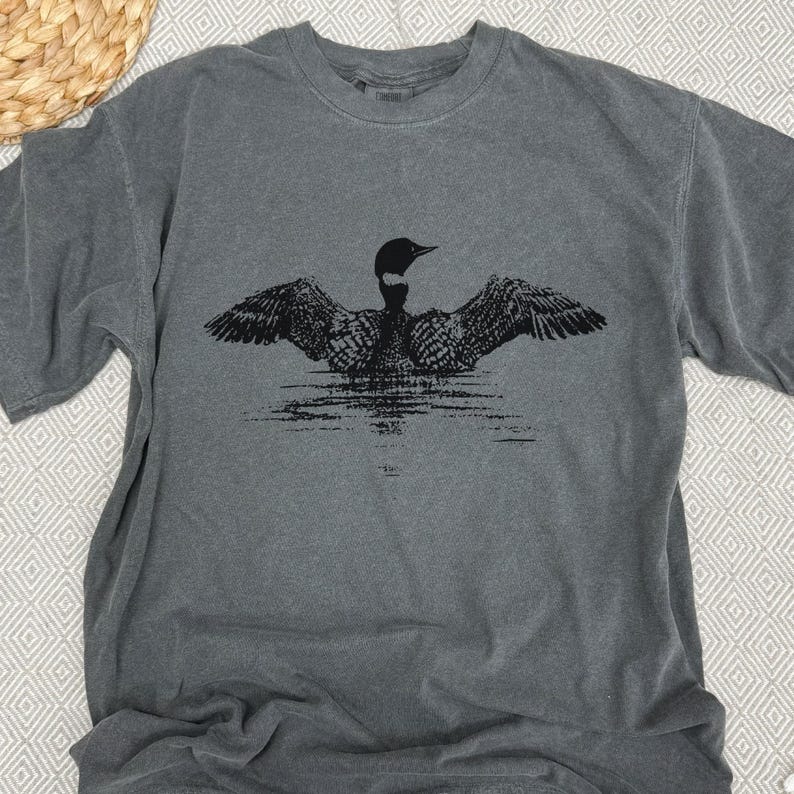 May include: A gray t-shirt featuring a black graphic of a loon with outstretched wings, appearing to float on water. The short-sleeved shirt has a crew neck and a slightly faded appearance, suggesting a vintage or washed style. The design is centered on the chest.