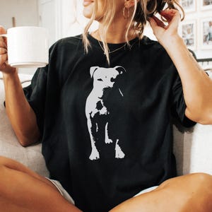 Pode incluir: Camiseta preta com um gr&aacute;fico branco de um cachorro. O cachorro &eacute; retratado em um estilo minimalista. A camiseta tem um ajuste folgado e grande. A imagem mostra um estilo casual.