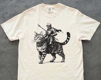 May include: Cream-colored t-shirt featuring a black and white illustration of a knight in full armor riding a large tabby cat. The knight holds a spear, and the cat has a striped tail. The shirt has a crew neck and short sleeves.