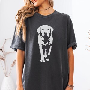 May include: Dark gray t-shirt featuring a white graphic of a Labrador Retriever dog. The dog is depicted in a walking pose, with a detailed outline. The shirt has short sleeves and a crew neck.