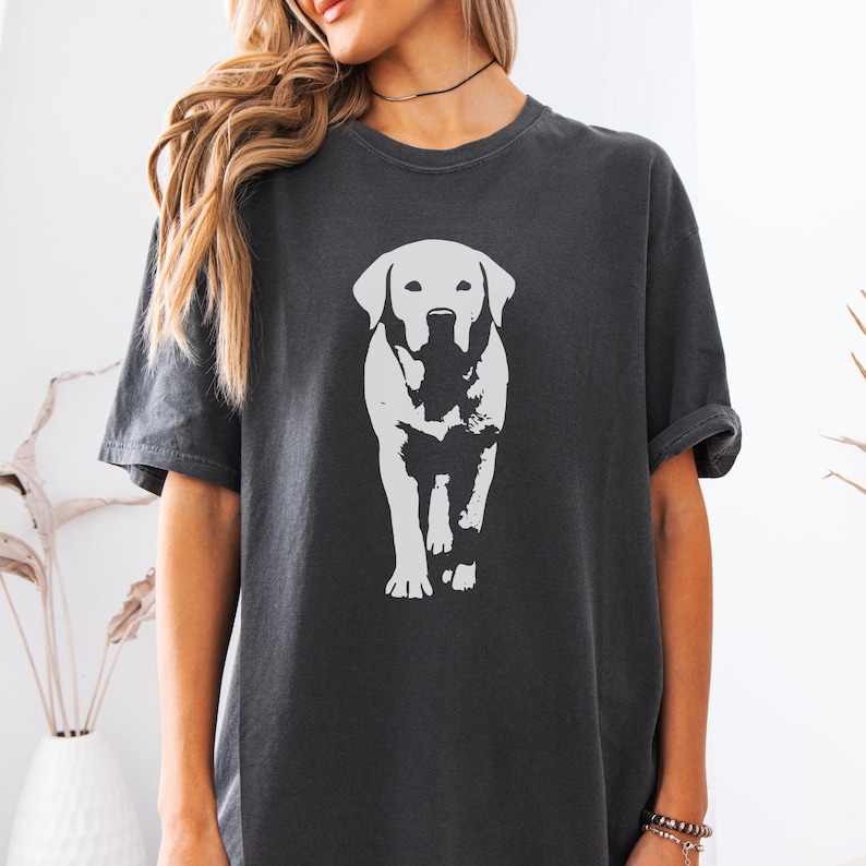 May include: Dark gray t-shirt featuring a white graphic of a Labrador Retriever dog. The dog is depicted in a walking pose, with a detailed outline. The shirt has short sleeves and a crew neck.