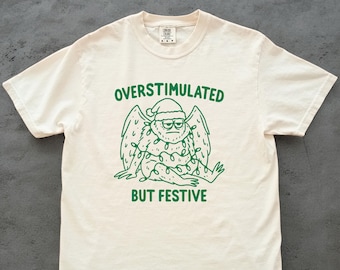Op de afbeelding: Crèmekleurig T-shirt met een groene afbeelding van een chagrijnig, gevleugeld wezen met een kerstmuts en kerstverlichting. De tekst "OVERSTIMULATED BUT FESTIVE" staat boven en onder de illustratie.