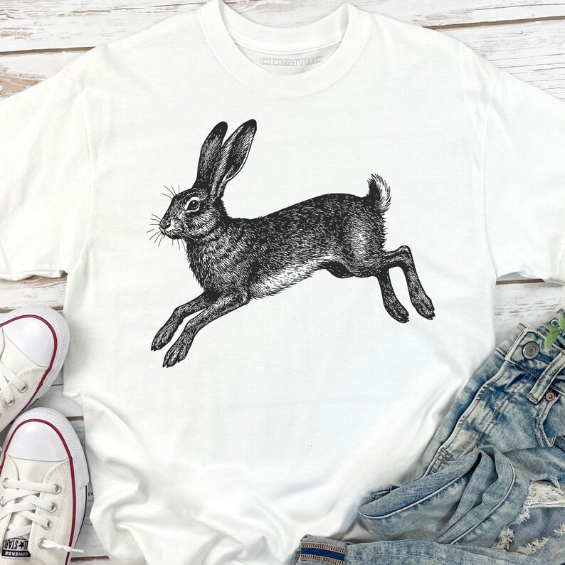 May include: White t-shirt featuring a detailed black and white illustration of a running hare. The design showcases the hare in mid-leap, with its ears erect and legs extended. The shirt is paired with denim jeans and white sneakers.